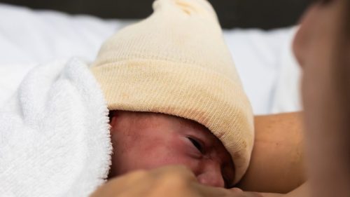 Newborn in white hat lies on person's chest in hospital setting.