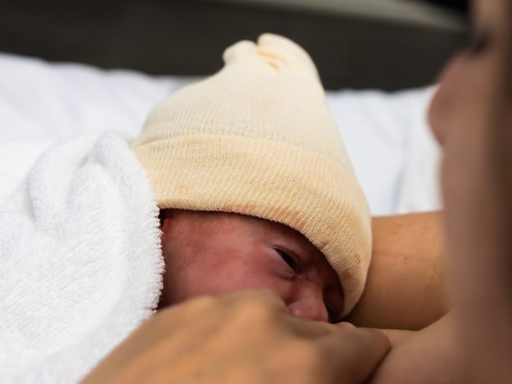 Newborn in white hat lies on person's chest in hospital setting.