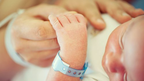Infant's hand grasping adult finger, hospital bracelet visible, evokes vulnerability and care.