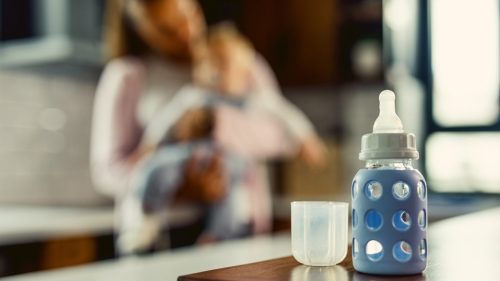 Blue baby bottle on counter, woman holding child blurred in background.