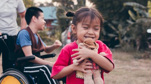 Smiling girl embracing teddy bear outside with wheelchair user in background.