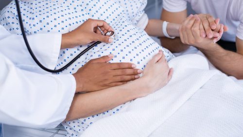 Doctor examines pregnant woman's belly while partner holds her hand.