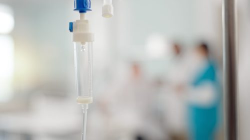 IV drip in hospital setting, with blurred healthcare professionals in background.