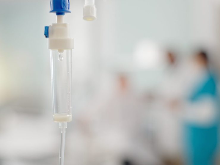 IV drip in hospital setting, with blurred healthcare professionals in background.