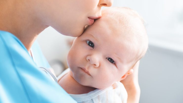 Woman in medical attire tenderly kissing baby's forehead.