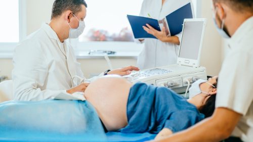 Doctor conducting ultrasound exam on pregnant woman, medical staff reviewing notes nearby.