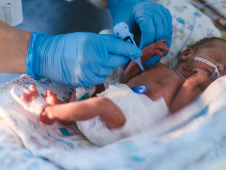 Premature infant receiving medical care in hospital incubator, monitored by gloved healthcare professional.